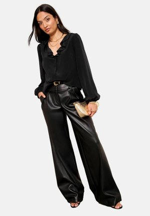 Schwarze, rüschige Bluse mit einer durchgehenden Knopfleiste, kombiniert mit hochgeschnittenen, weit geschnittenen schwarzen Kunstlederhosen und einer kleinen goldenen Handtasche.