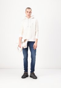 Young man wearing a white patterned hoodie, blue jeans, dark sneakers, holding a beige tote bag, standing against a white background.