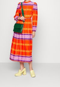 Robe midi à motifs rouges et orange avec des rayures violettes et jaunes, associée à un sac à main en crocodile vert et des chaussures à talons block jaunes.