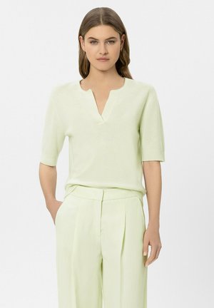 Woman wearing light green short-sleeve sweater with notch neckline and matching high-waist pleated trousers standing with one hand in pocket.