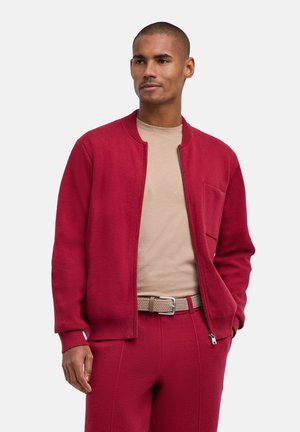 Uomo che indossa una giacca rossa con zip e pantaloni abbinati con una camicia beige e cintura intrecciata, in piedi con una mano in tasca.