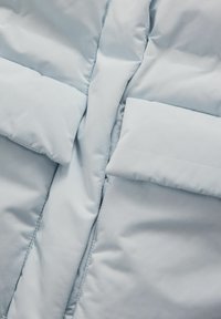 Lyseblå pufferjakke med en glat tekstur, der har et quiltet design og to frontlommer. Lynlås lukning i midten.