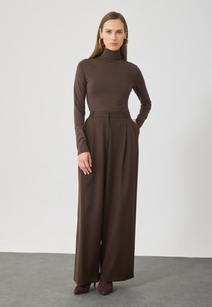 Maglione a collo alto marrone e pantaloni marroni a vita alta e a gamba larga. Il tessuto sembra morbido con una consistenza liscia. Le scarpe con tacco sono di un bordeaux scuro.