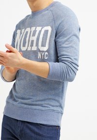 Blue sweatshirt with white "NOHO NYC" text. Features ribbed cuffs and hem, made of soft, textured fabric in a casual design.