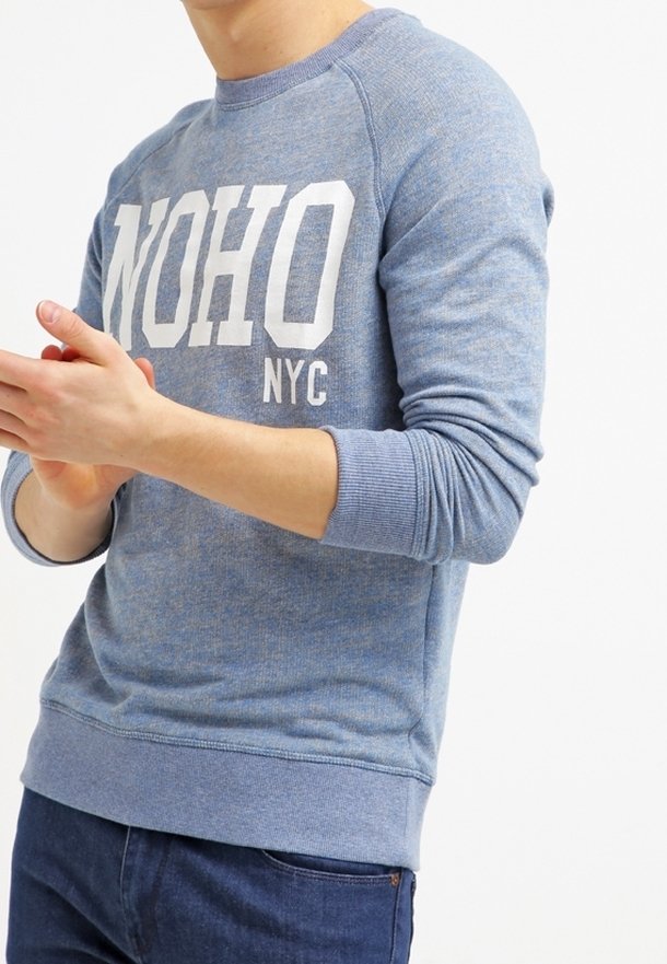 Blue sweatshirt with white "NOHO NYC" text. Features ribbed cuffs and hem, made of soft, textured fabric in a casual design.