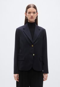 Marineblauwe blazer met een getailleerde pasvorm, klepzakken en twee gouden knopen. Heeft revers met inkeping en een gladde textuur, gecombineerd met een zwarte coltrui.