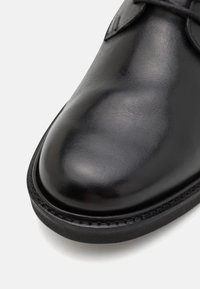 Bout de chaussure habillée en cuir noir poli avec détail à lacets et semelle cousue sur fond blanc.
