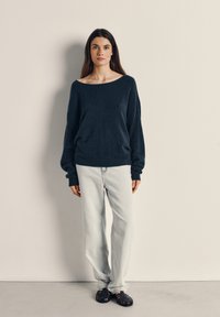 Marineblå oversize striktrøje med off-shoulder halsudskæring, parret med lyse jeans med lige ben og sorte sandaler med strappedesign.