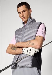 Sivi puffer prsluk s horizontalnim šavovima, uparen s ružičastom polo majicom. Outfit uključuje golf štap i bijelu golf rukavicu s crnim detaljima.