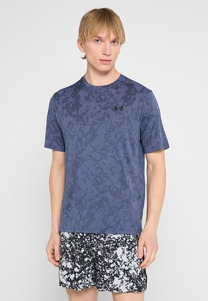 Młody mężczyzna w niebieskiej koszulce Under Armour z wzorem i czarno-białych szortach z abstrakcyjnym nadrukiem, stojący na jednolitym tle.