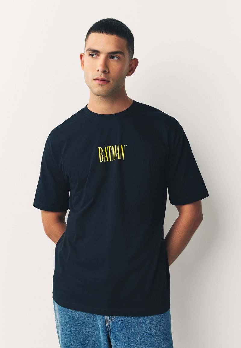 Camiseta negra de algodón con un logotipo amarillo de "BATMAN" centrado en el pecho, mangas cortas y un cuello redondo clásico.