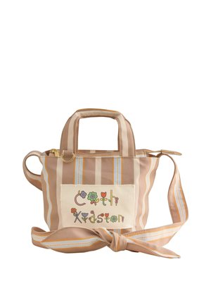 Ung kvinde iført en beige top, lyse, stribede bukser og en stribet crossbody taske med farverig tekst på en blå væg.