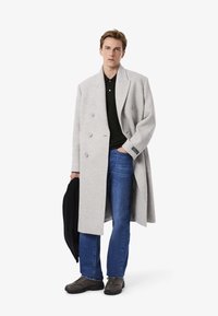 Cappotto in lana grigio doppiopetto con grandi bottoni, abbinato a una polo nera, jeans blu e sneakers scure. Il cappotto presenta un design oversize.