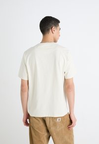 T-shirt en coton crème à manches courtes et col rond, porté par un modèle vêtu de pantalons en velours côtelé. Texture subtile, pas de graphismes visibles.
