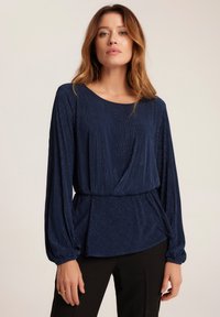 Granatowa bluzka z długimi rękawami i marszczoną talią. Posiada teksturowany materiał z pionowymi paskami oraz okrągłym dekoltem.