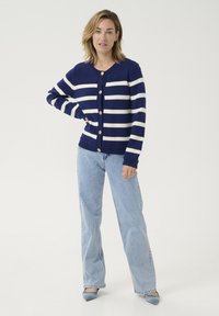 Navy blue cardigan with white stripes, featuring wooden buttons and a ribbed hem. Paired with light blue jeans and blue pointed shoes.