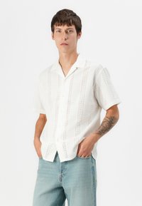 Camisa blanca de manga corta con una tela texturizada que presenta patrones de cortes verticales. Estilo abotonado y corte relajado.