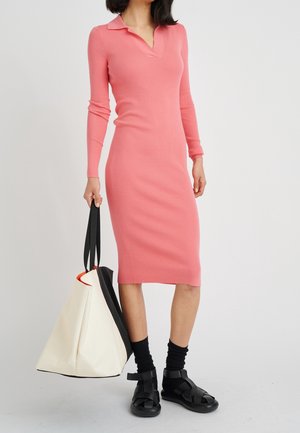 Femme portant une robe midi côtelée rose à manches longues, des chaussettes noires, des sandales noires, tenant un grand sac cabas blanc et noir.