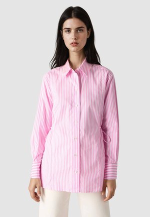 Kobieta ubrana w koszulę na guziki z pionowymi różowo-białymi paskami z długim rękawem oraz białe spodnie na jednolitym jasnym tle.