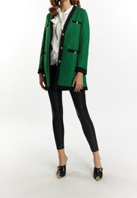 Blazer verde texturizado con ribete negro y botones dorados, combinado con leggings de cuero negro y mules negras con detalles dorados.