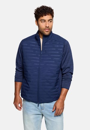 Hombre con cabello rizado que lleva un chaleco azul marino con cremallera sobre una camisa de manga larga y jeans azul claro, mirando ligeramente hacia la derecha.