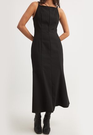 Kobieta w czarnej, bezrękawnikowej sukience midi z pionowymi szwami i kwadratowym dekoltem, zestawiona z czarnymi botkami na obcasie.