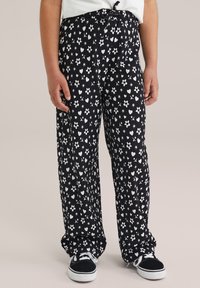Black pants with a floral and heart pattern in white. Made of soft fabric, featuring an elastic waistband and loose fit. Worn with black sneakers.