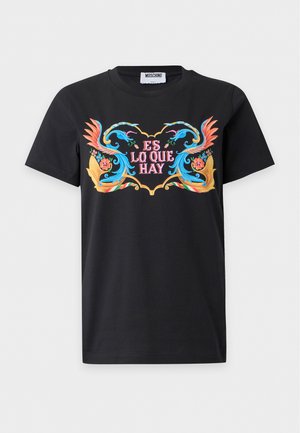 Femme debout, les mains dans les poches, portant un t-shirt noir avec un motif graphique, un pantalon gris avec une ceinture noire et des sandales rouges sur fond blanc.