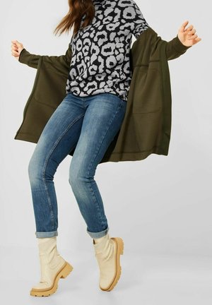 Personne portant un pull à motif léopard, un jean bleu retroussé, des bottes couleur crème et une veste olive ouverte, capturée en mouvement sur un fond uni.