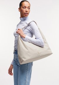 Sac à épaule en nylon beige avec une texture lisse, une forme spacieuse et des accessoires en métal argenté ; présente un logo discret sur le côté.