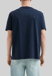 Granatowa bawełniana koszulka z krótkim rękawem, z ukośnym detalem na plecach i luźnym krojem. Noszona z jasnoniebieskimi dżinsami.