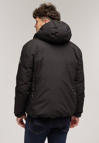 Czarna kurtka puchowa z dużym kapturem, pikowaną fakturą i elastycznymi mankietami. Posiada naszywkę z logo na rękawie. Nosi się ją na ciemnych dżinsach.