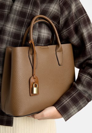 Brązowa torebka z teksturowanej skóry z złotą kłódką trzymana przez osobę w ciemnobrązowej kratowanej marynarce i kremowej prążkowanej spódnicy.