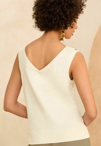Cream-colored sleeveless top with a V-neck and ribbed hem. Notable details include button accents at the shoulder and textured fabric.