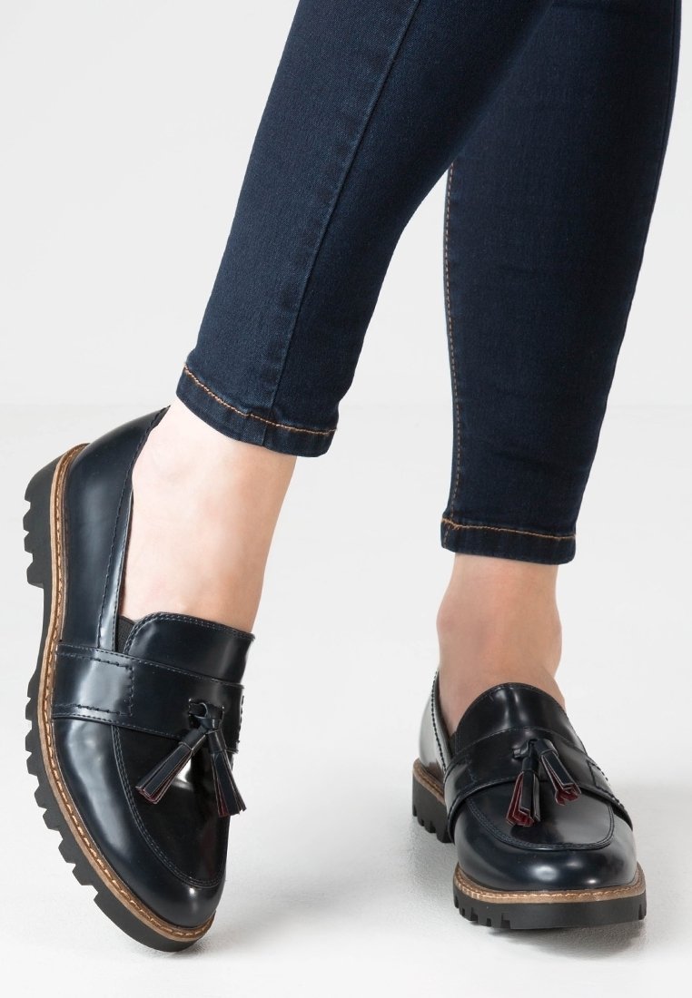 Mocassins en cuir noir à pampilles portés avec des jeans skinny bleu foncé sur un fond blanc.
