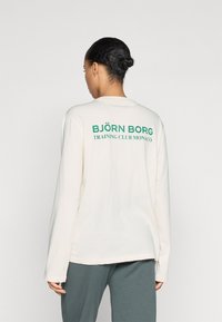 Long-sleeve cream shirt featuring green text "BJÖRN BORG TRAINING CLUB MONACO" on the back. Soft fabric with a relaxed fit.