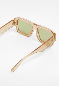 Transparent light brown sunglasses with square frames, green lenses, and a clean design. Features a visible brand engraving on the temple.