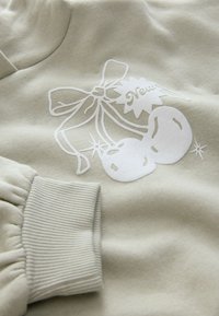 Világos zöld pulóver fehér cseresznye grafikával, masnival és az "Új" szóval. Mandzsettás ujj bordázott textúrával. Puha anyagból.