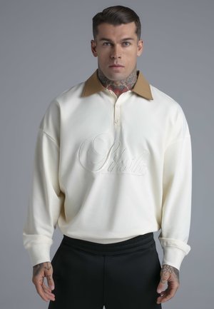 Mężczyzna z tatuażami na szyi i dłoni, ubrany w białą koszulę z długim rękawem i brązowym kołnierzem oraz haftem "Siksilk" z przodu.