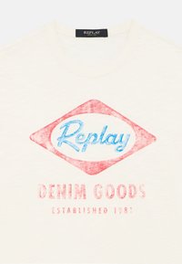 Camiseta de algodón color crema con un diseño retro que presenta una forma de diamante roja y el logo azul de "Replay", junto con el texto "DENIM GOODS" en rojo desvanecido.