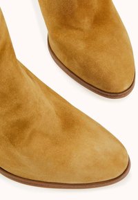 Bottes cheville en daim beige avec bouts arrondis et semelles en cuir marron sur fond blanc.