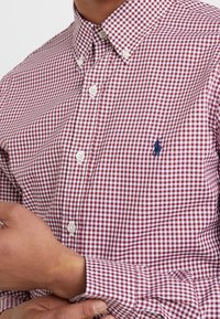 Nära bild av en person som bär en röd- och vitrutig skjorta med knappar, med en liten marinblå broderad logotyp på bröstet.