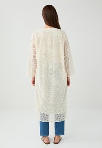 Langer, offener Spitzenkimono in Off-White mit geometrischen Mustern. Verfügt über weite Ärmel und eine lockere Passform, getragen über blauen Jeans.