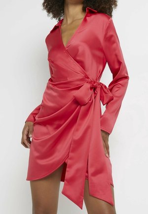 Femme portant une robe portefeuille rouge brillante à manches longues avec un grand nœud latéral noué à la taille, debout devant un fond blanc.