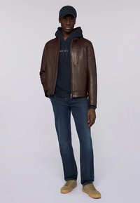 Brun läderjacka med dragkedja, marinblå huvtröja under, mörkblå jeans och beiga sneakers. Svart keps på huvudet.