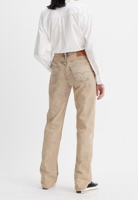 Jean en denim beige avec une coupe droite, présentant des poches arrière et une étiquette en cuir de marque à la taille. Porté avec une chemise blanche.