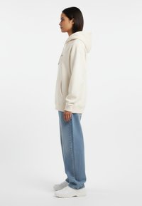 Felpa crema con grande tasca frontale, cappuccio con cordoncino e logo ricamato, abbinata a jeans a gamba larga blu chiaro e sneakers bianche.