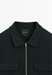 Veste noire à col, fabriquée en tissu texturé, avec une fermeture éclair sur le devant et deux poches poitrine, arborant une étiquette avec la mention "Massimo Dutti."