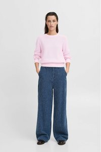 Helle rosa, strukturierte Strickjacke mit rundem Ausschnitt, kombiniert mit weit geschnittenen blauen Cordhosen mit vertikalen Streifen. Braune Schuhe runden das Outfit ab.