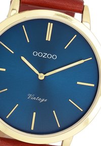 Montre dorée avec un cadran bleu mat, des index et des aiguilles dorés. Équipée d'un bracelet en cuir brun et du texte "Vintage" près du bas.